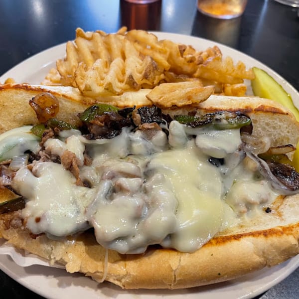 Cheesesteak sandwich with melted cheese, peppers, onions, and waffle fries on a white plate.