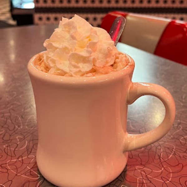 A RedEye Diner hot chocolate with whipped cream and a spoon, on a patterned diner table.